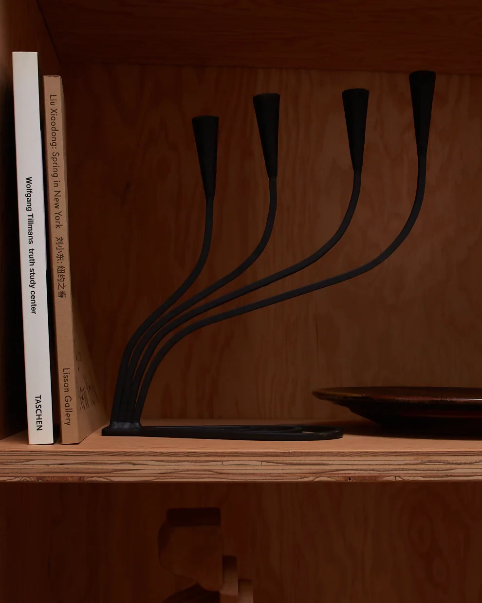 A black, hand forged iron candle holder with four curved arms and cone shaped heads, positioned on a wooden shelf next to a book,  available  to buy on the-salon.com. SALON curates the finest , unique, handmade pieces from the interior industry's best makers and brands. Shop luxury furniture, lighting, rugs, art, antiques and vintage pieces and accessories from SALON interior designed rooms.
