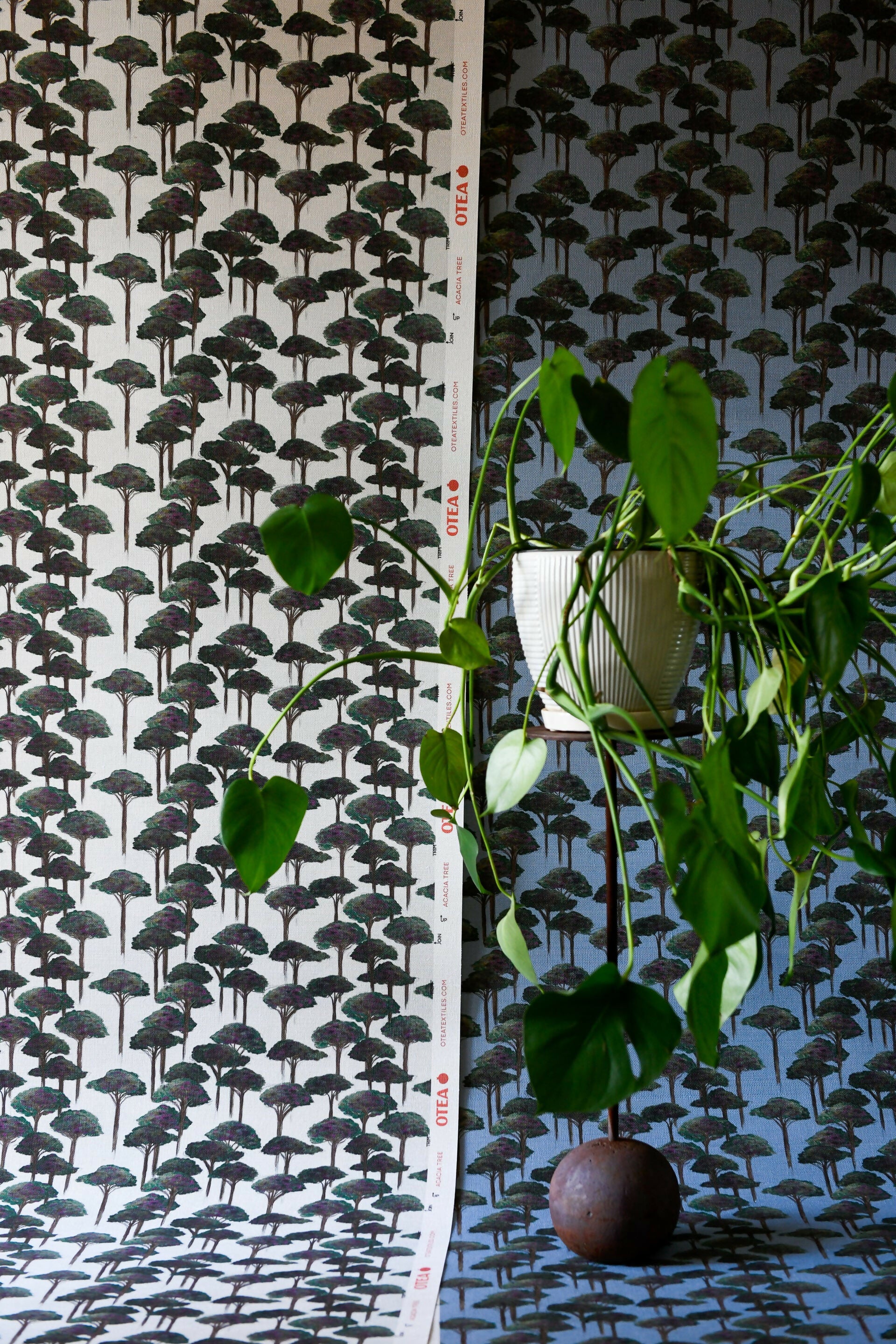 A room with wallpaper featuring a pattern of stylized pine trees and umbrellas in green shades, with a plant in the foreground, available  to buy on the-salon.com. SALON curates the finest , unique, handmade pieces from the interior industry's best makers and brands. Shop luxury furniture, lighting, rugs, art, antiques and vintage pieces and accessories from SALON interior designed rooms.