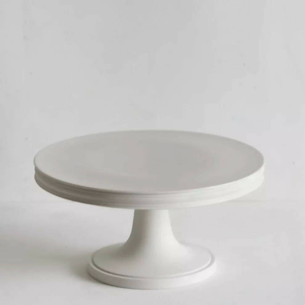 A white porcelain cake stand with a classic design, featuring a round platform on a cylindrical pedestal,  available  to buy on the-salon.com. SALON curates the finest , unique, handmade pieces from the interior industry's best makers and brands. Shop luxury furniture, lighting, rugs, art, antiques and vintage pieces and accessories from SALON interior designed rooms.