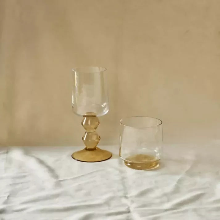 Two glass tumblers with gold bases on a light fabric surface,  available  to buy on the-salon.com. SALON curates the finest , unique, handmade pieces from the interior industry's best makers and brands. Shop luxury furniture, lighting, rugs, art, antiques and vintage pieces and accessories from SALON interior designed rooms.
