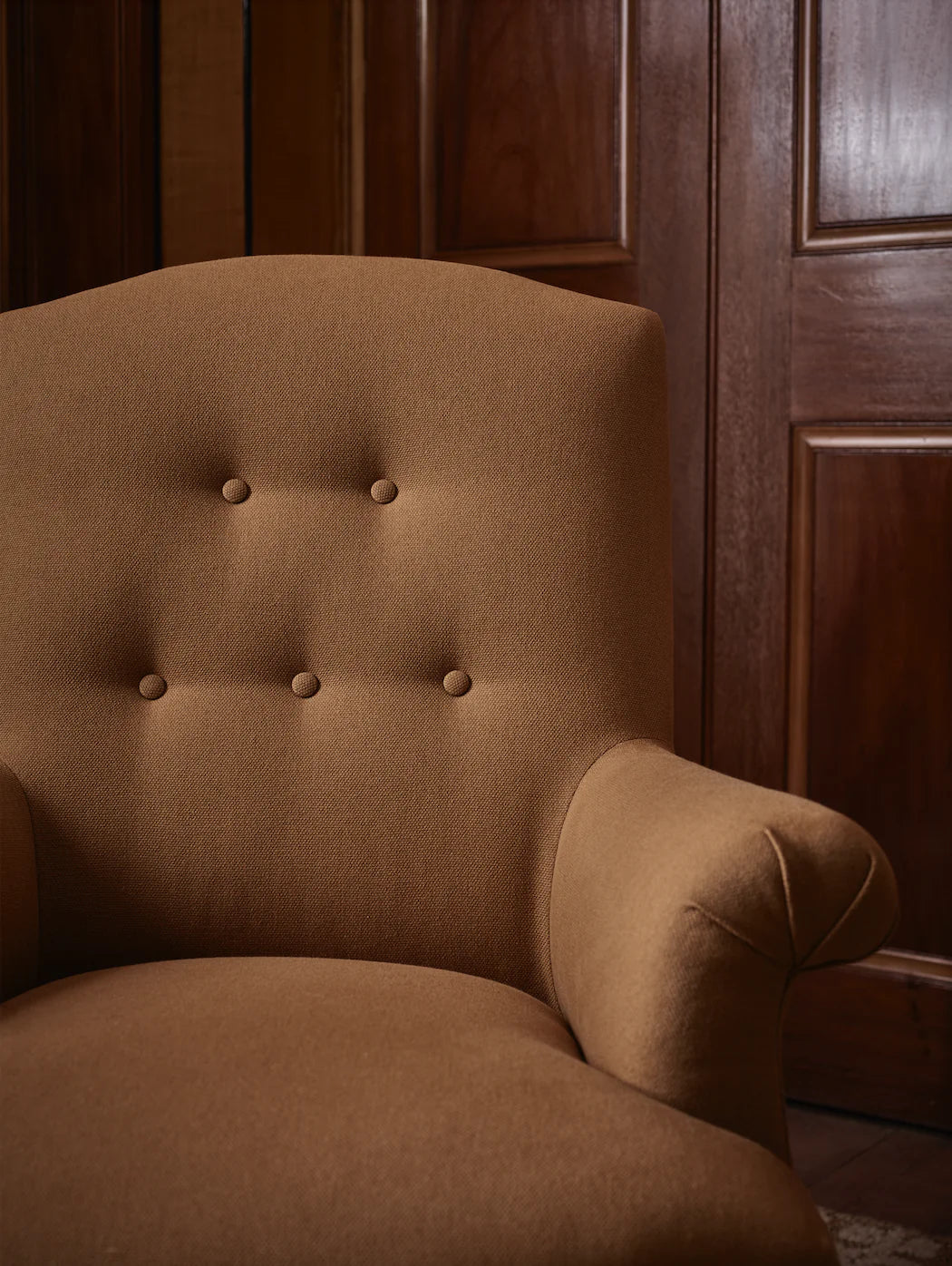 A brown upholstered armchair with button back detailing and scroll arms, set against a wooden panel background,  available  to buy on the-salon.com. SALON curates the finest , unique, handmade pieces from the interior industry's best makers and brands. Shop luxury furniture, lighting, rugs, art, antiques and vintage pieces and accessories from SALON interior designed rooms.