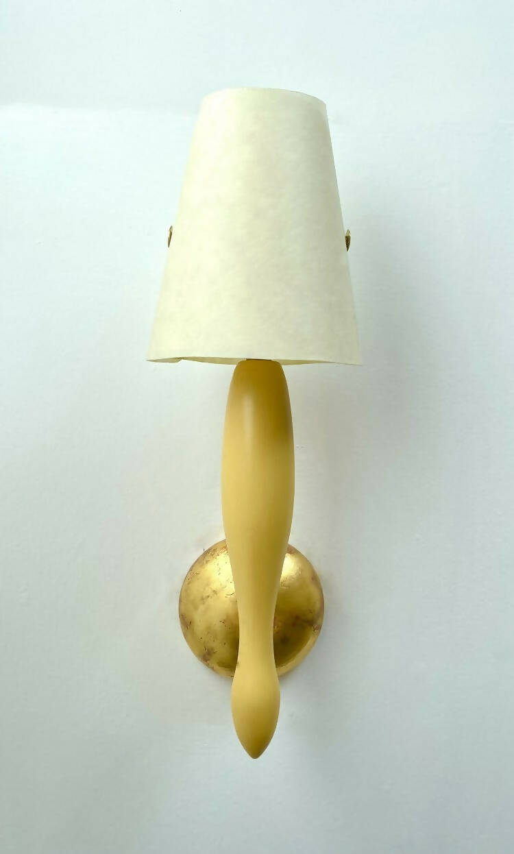A wall light fixture with a yellow, stem-like structure and a white conical shade, mounted on a gold-colored plate, available  to buy on the-salon.com. SALON curates the finest , unique, handmade pieces from the interior industry's best makers and brands. Shop luxury furniture, lighting, rugs, art, antiques and vintage pieces and accessories from SALON interior designed rooms.