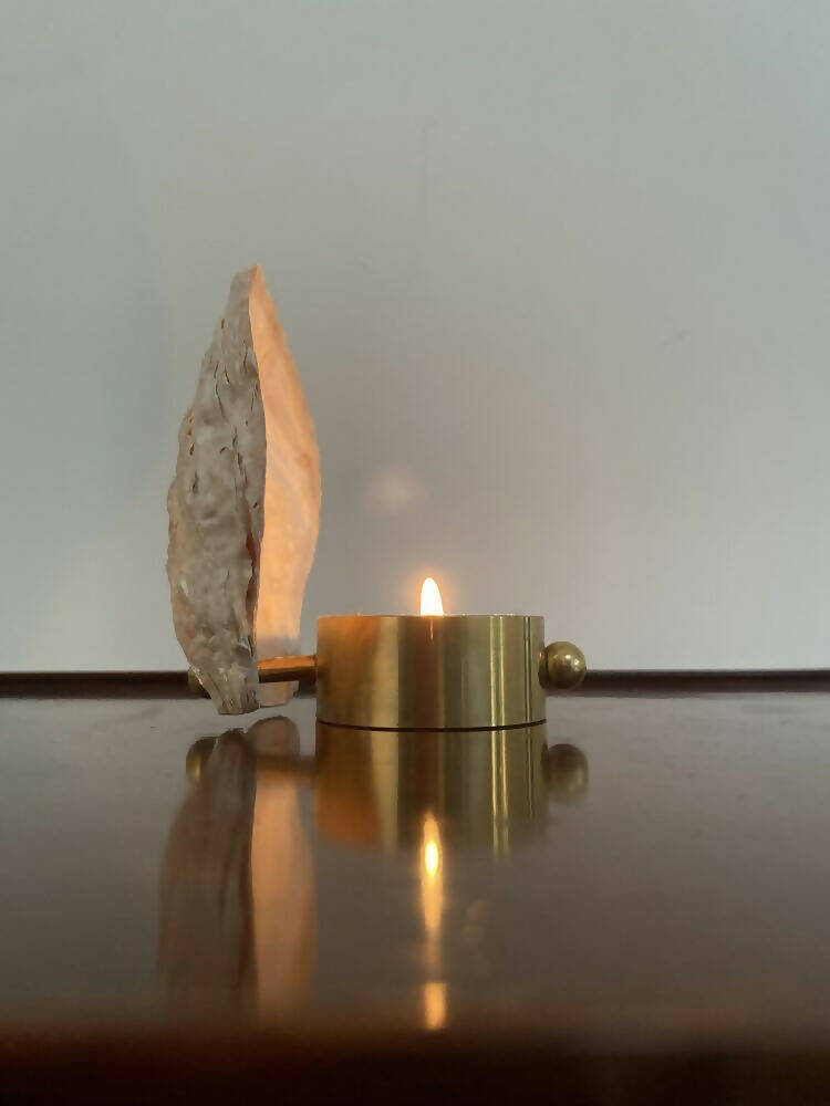 A brass tealight candle holder in the shape of a Norfolk oyster shell, with a lit candle,  displayed against a neutral background,  available  to buy on the-salon.com. SALON curates the finest , unique, handmade pieces from the interior industry's best makers and brands. Shop luxury furniture, lighting, rugs, art, antiques and vintage pieces and accessories from SALON interior designed rooms.