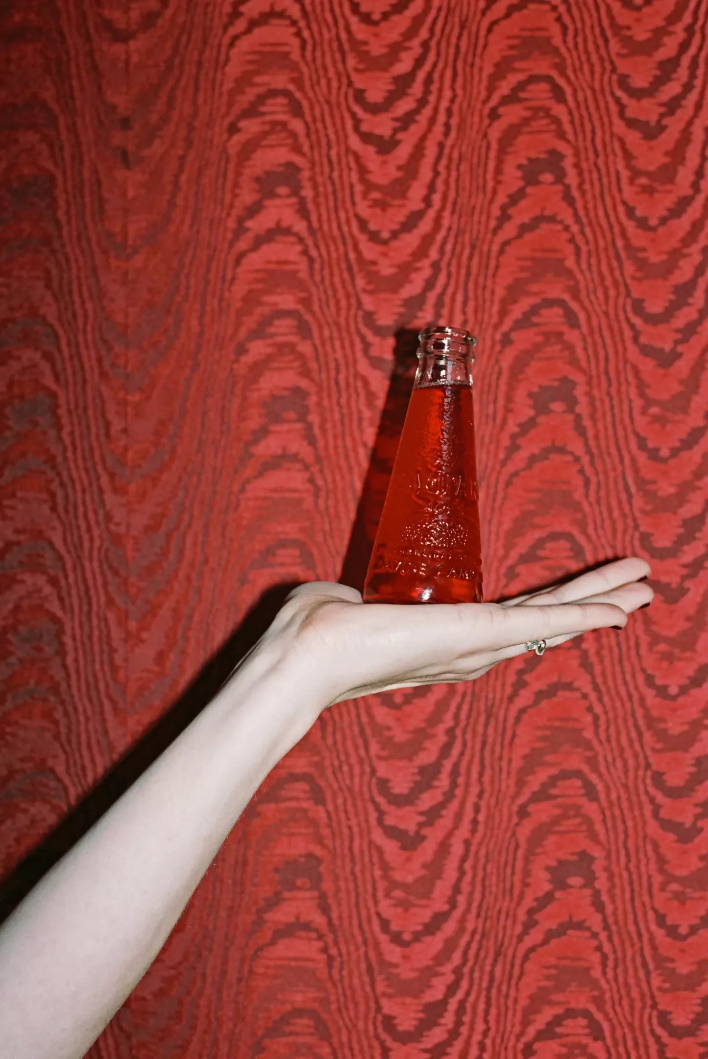 An image of a red moiré patterned fabric on the walls, behind a glass with red liquid and someone holding it,  available  to buy on the-salon.com. SALON curates the finest , unique, handmade pieces from the interior industry's best makers and brands. Shop luxury furniture, lighting, rugs, art, antiques and vintage pieces and accessories from SALON interior designed rooms.