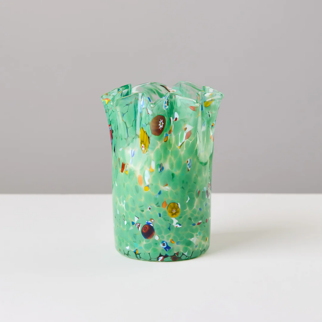 A custom hand-blown green glass candle holder with embedded colorful glass pieces, displayed alongside its lid, on a white surface,  available  to buy on the-salon.com. SALON curates the finest , unique, handmade pieces from the interior industry's best makers and brands. Shop luxury furniture, lighting, rugs, art, antiques and vintage pieces and accessories from SALON interior designed rooms.
