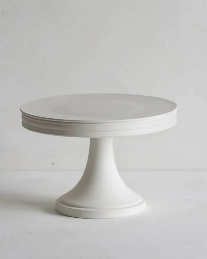 A white porcelain cake stand with a simple, elegant design featuring a flat platform on top and a cylindrical base, available  to buy on the-salon.com. SALON curates the finest , unique, handmade pieces from the interior industry's best makers and brands. Shop luxury furniture, lighting, rugs, art, antiques and vintage pieces and accessories from SALON interior designed rooms.