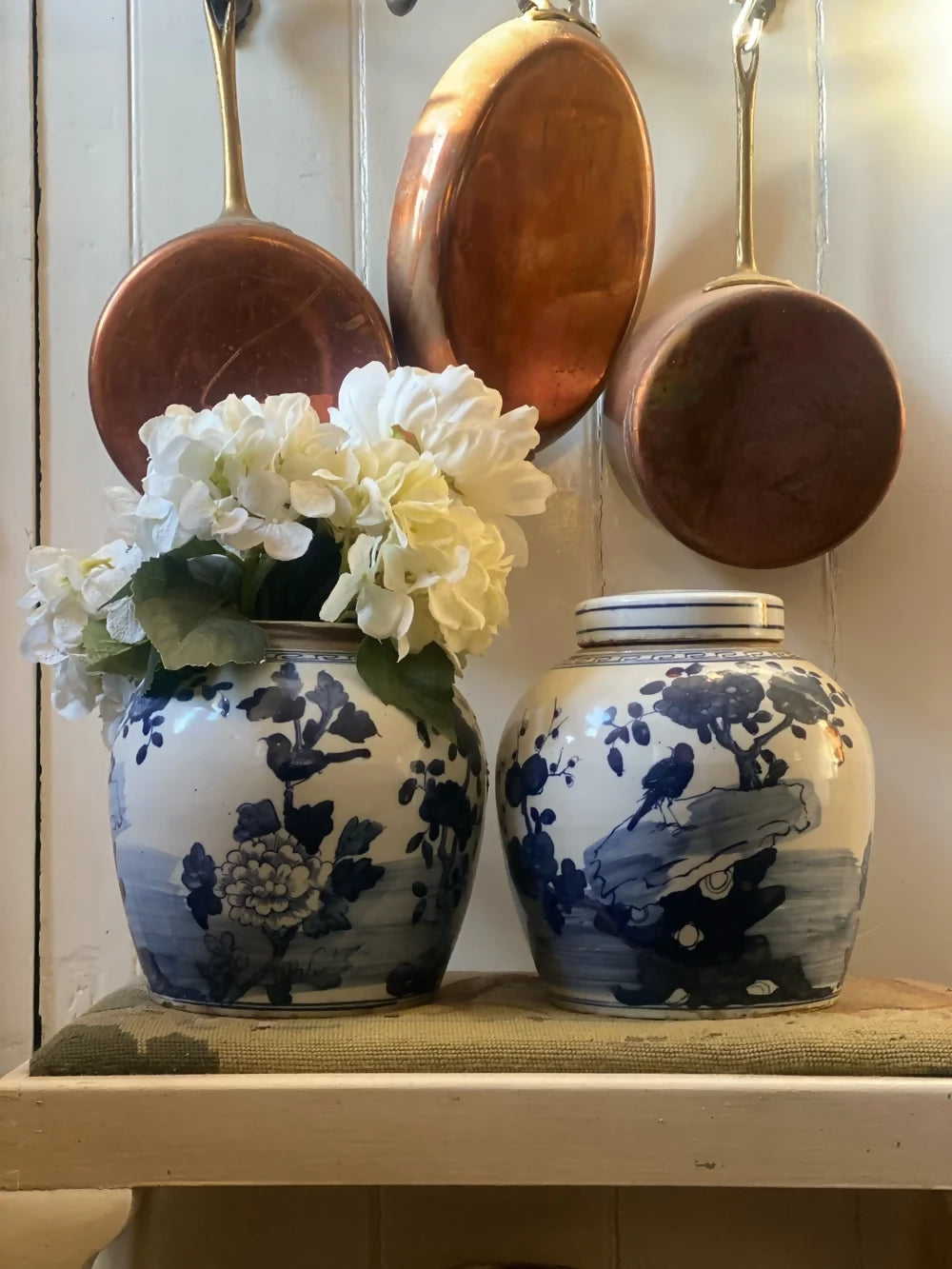 Two blue and white ceramic jars with floral designs on a shelf, with copper pots hanging above,  available  to buy on the-salon.com. SALON curates the finest , unique, handmade pieces from the interior industry's best makers and brands. Shop luxury furniture, lighting, rugs, art, antiques and vintage pieces and accessories from SALON interior designed rooms.