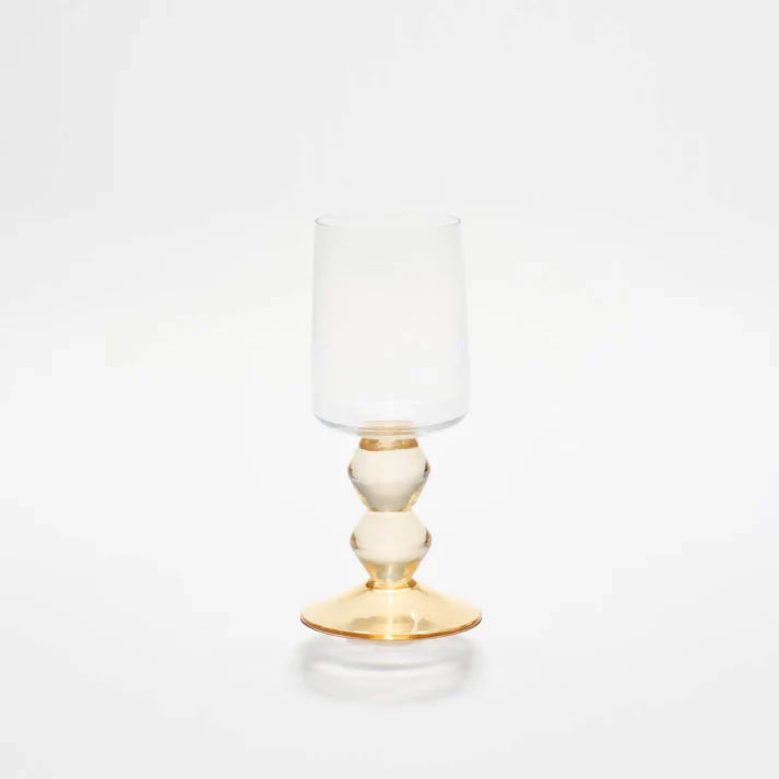 A clear wine glass with a gold-stemmed base and a spherical shape, with a clear glass body and a round, white surface at the top, available  to buy on the-salon.com. SALON curates the finest , unique, handmade pieces from the interior industry's best makers and brands. Shop luxury furniture, lighting, rugs, art, antiques and vintage pieces and accessories from SALON interior designed rooms.