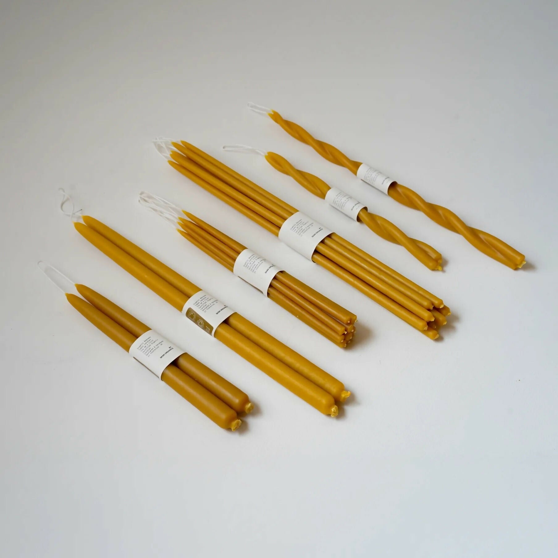 A group of unlit organic beeswax dinner candles, in various shapes including straight and twisted, hanging from a metal rod,  available  to buy on the-salon.com. SALON curates the finest , unique, handmade pieces from the interior industry's best makers and brands. Shop luxury furniture, lighting, rugs, art, antiques and vintage pieces and accessories from SALON interior designed rooms.