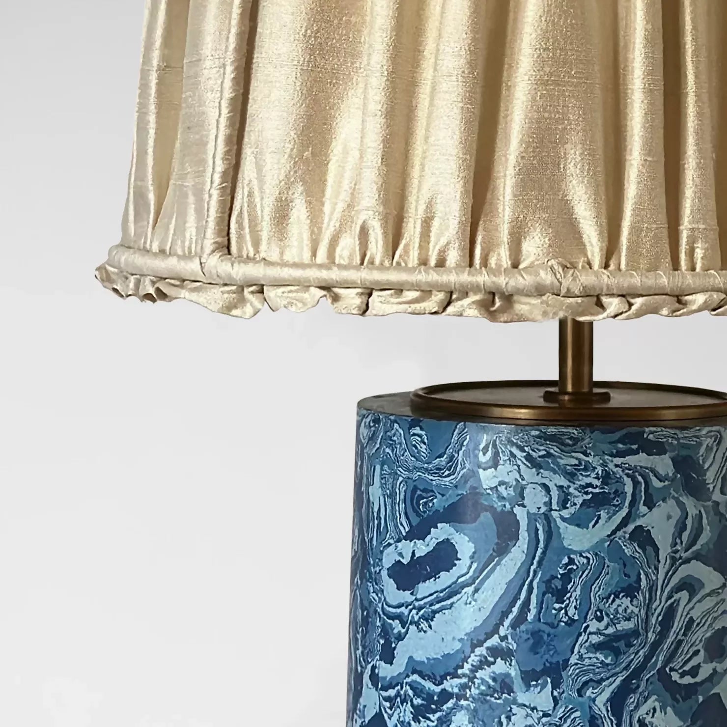 Close up of a  blue marbled base table lamp with brass detailing, topped with a gold pleated shade,  available  to buy on the-salon.com. SALON curates the finest , unique, handmade pieces from the interior industry's best makers and brands. Shop luxury furniture, lighting, rugs, art, antiques and vintage pieces and accessories from SALON interior designed rooms.