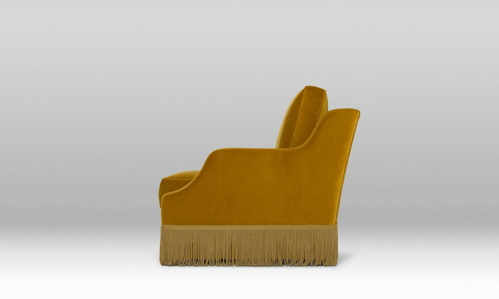 The side of a Mustard yellow armchair with fringe on a light gray background, available  to buy on the-salon.com. SALON curates the finest , unique, handmade pieces from the interior industry's best makers and brands. Shop luxury furniture, lighting, rugs, art, antiques and vintage pieces and accessories from SALON interior designed rooms.