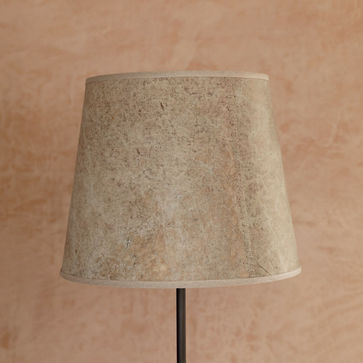 A beige hand stitched tapered lampshade made of Lokta paper with a natural linen trim, displayed on a lamp stand against a pink background,  available  to buy on the-salon.com. SALON curates the finest , unique, handmade pieces from the interior industry's best makers and brands. Shop luxury furniture, lighting, rugs, art, antiques and vintage pieces and accessories from SALON interior designed rooms.