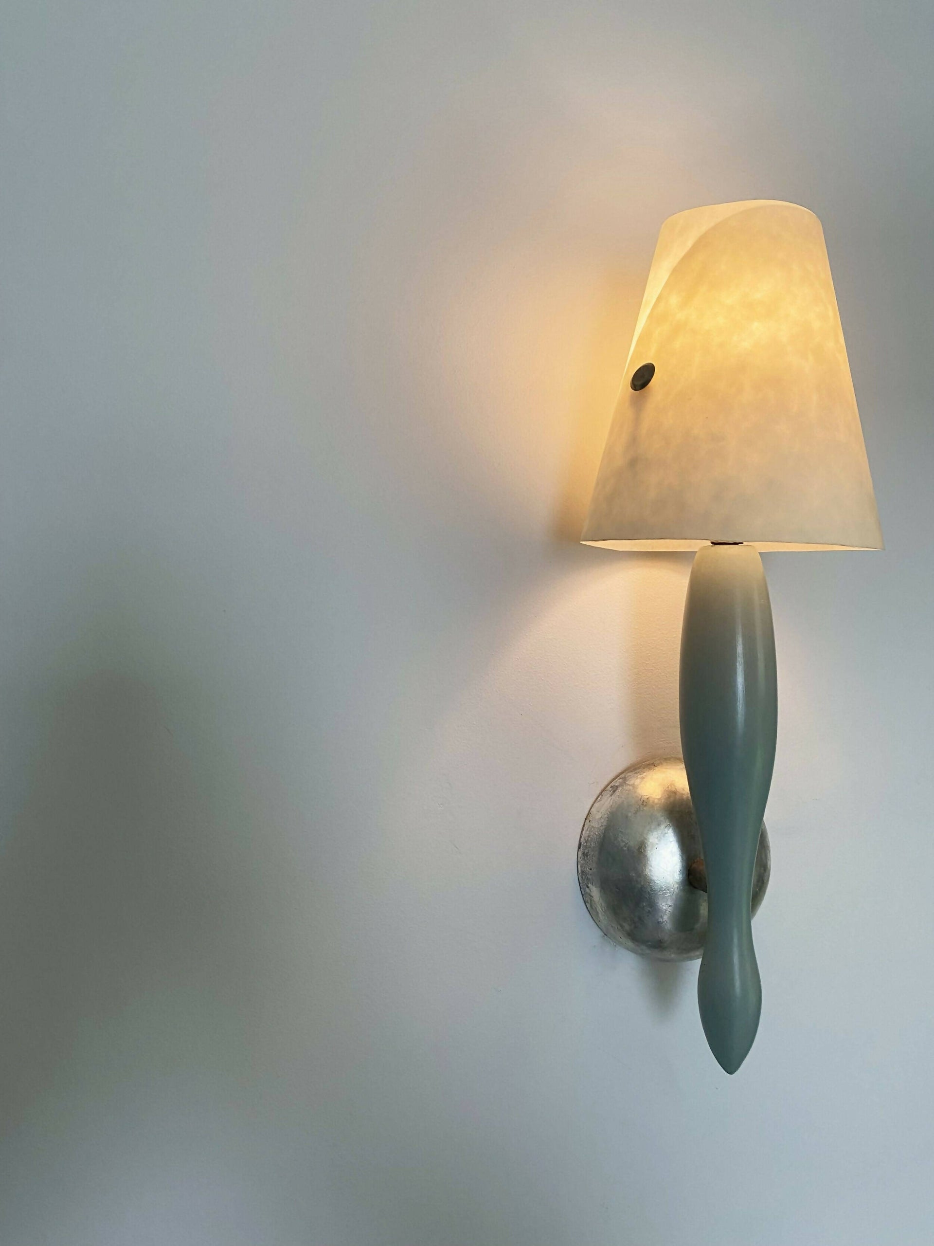 A wall light fixture with a blue, stem-like structure and a white conical shade, mounted on a silver-colored plate, available  to buy on the-salon.com. SALON curates the finest , unique, handmade pieces from the interior industry's best makers and brands. Shop luxury furniture, lighting, rugs, art, antiques and vintage pieces and accessories from SALON interior designed rooms.