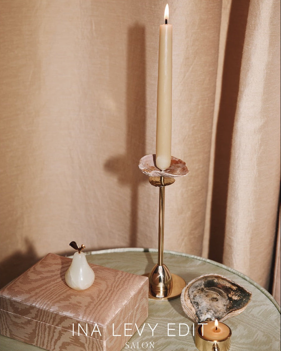 Candle in a gold holder on a table with a wooden box and decorative items, featuring 'INA LEVY EDIT' branding.