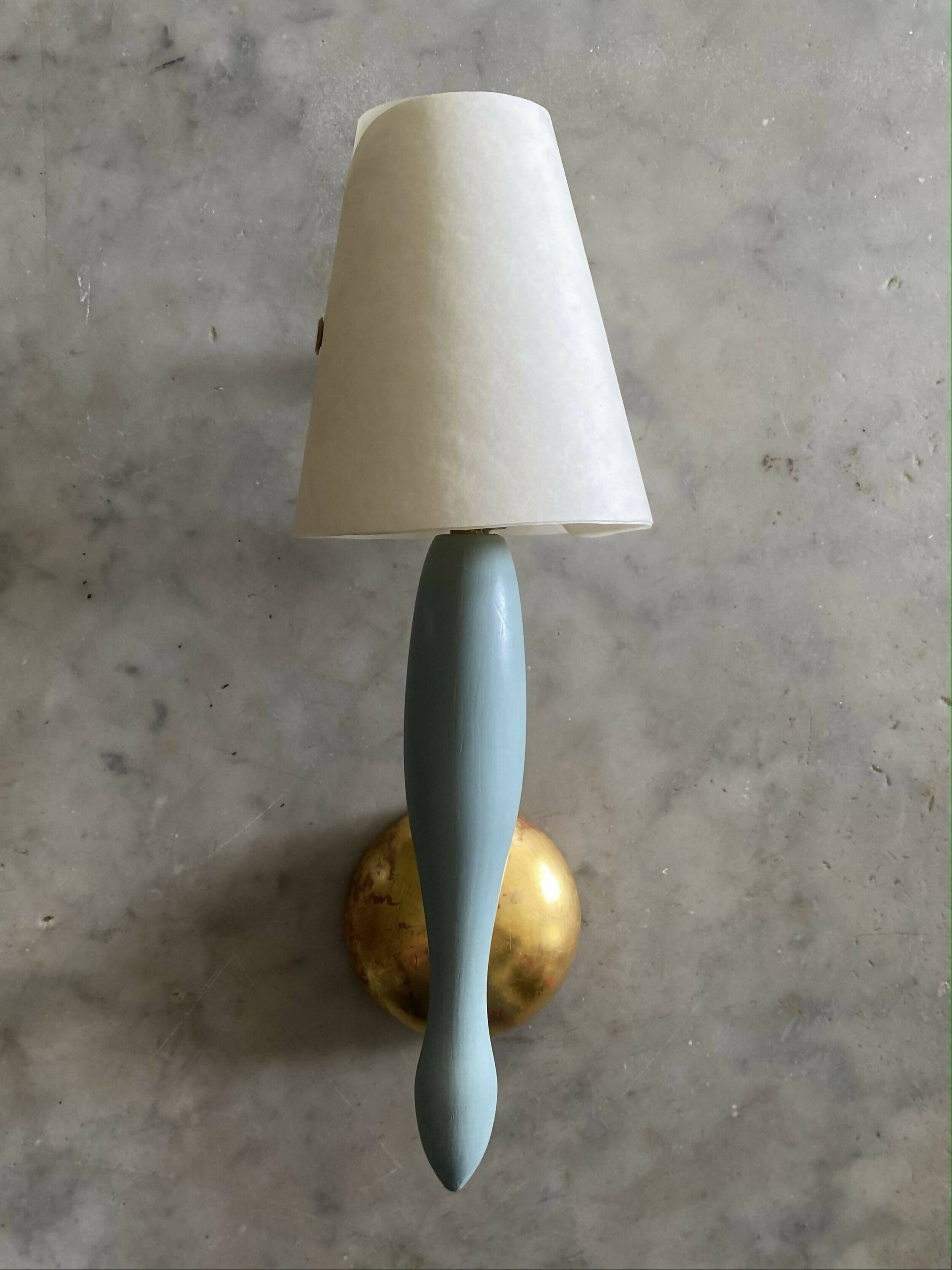 A wall light fixture with a blue, stem-like structure and a white conical shade, mounted on a gold-colored plate, available  to buy on the-salon.com. SALON curates the finest , unique, handmade pieces from the interior industry's best makers and brands. Shop luxury furniture, lighting, rugs, art, antiques and vintage pieces and accessories from SALON interior designed rooms.