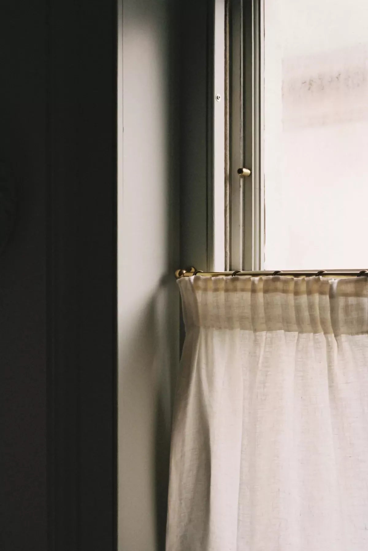 Window with a curtain partially drawn, showing a soft light on a dark wall, showing the curtain pole,  available  to buy on the-salon.com. SALON curates the finest , unique, handmade pieces from the interior industry's best makers and brands. Shop luxury furniture, lighting, rugs, art, antiques and vintage pieces and accessories from SALON interior designed rooms.