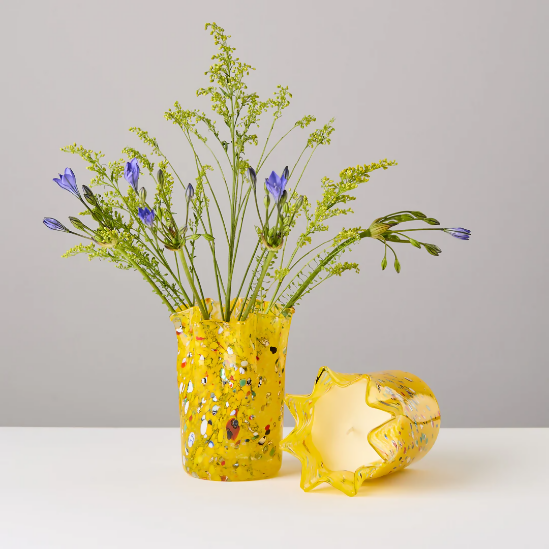 A custom hand-blown yellow candle holder with multicolor speckles, alongside a candle that has been melted to reveal the wax inside, placed on a white surface,  available  to buy on the-salon.com. SALON curates the finest , unique, handmade pieces from the interior industry's best makers and brands. Shop luxury furniture, lighting, rugs, art, antiques and vintage pieces and accessories from SALON interior designed rooms.