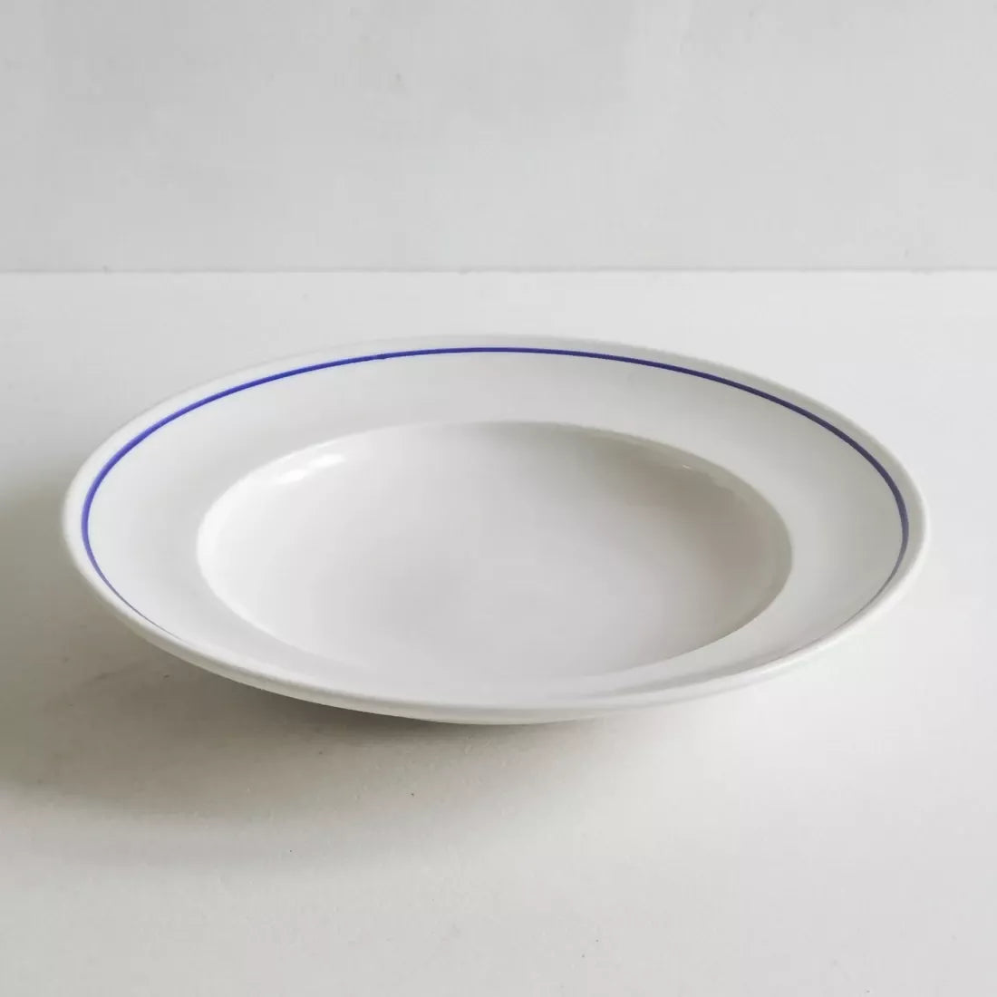 A white porcelain shallow bowl with a cobalt blue line along the rim, against a plain background, available  to buy on the-salon.com. SALON curates the finest , unique, handmade pieces from the interior industry's best makers and brands. Shop luxury furniture, lighting, rugs, art, antiques and vintage pieces and accessories from SALON interior designed rooms.