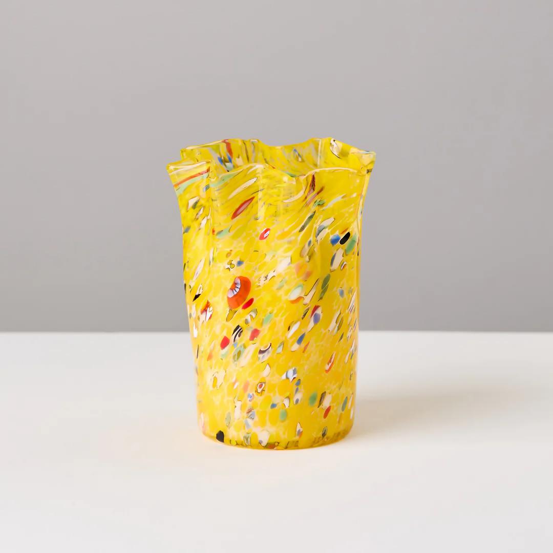 A custom hand-blown yellow candle holder with multicolor speckles, alongside a candle that has been melted to reveal the wax inside, placed on a white surface,  available  to buy on the-salon.com. SALON curates the finest , unique, handmade pieces from the interior industry's best makers and brands. Shop luxury furniture, lighting, rugs, art, antiques and vintage pieces and accessories from SALON interior designed rooms.