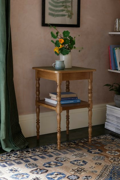 A brown wooden bedside table with a leather top, featuring a vase with flowers on top and a book leaning against it, placed on a patterned carpet,  available  to buy on the-salon.com. SALON curates the finest , unique, handmade pieces from the interior industry's best makers and brands. Shop luxury furniture, lighting, rugs, art, antiques and vintage pieces and accessories from SALON interior designed rooms.