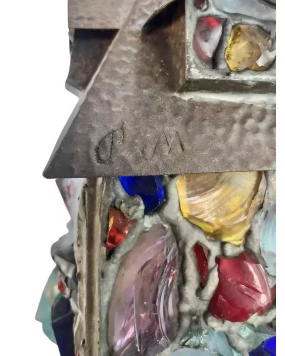 Close-up of a mixed-media art piece featuring a hammered metallic gray triangular element with an embossed monogram, set against a background of colorful glass inclusions in a textured resin or plaster-like material.