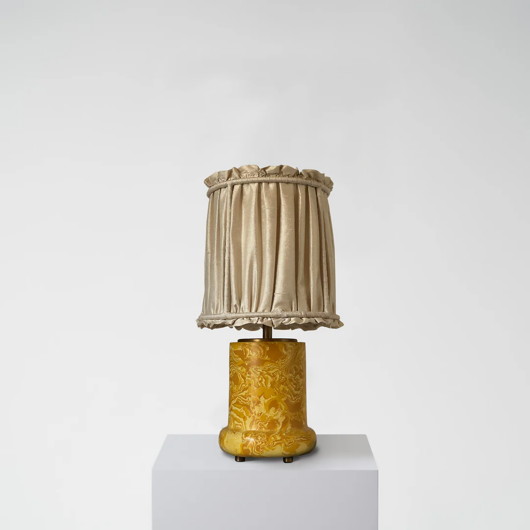 A yellow scagliola table lamp with a floral pattern base and a beige lampshade, displayed on a white platform against a neutral background, available  to buy on the-salon.com. SALON curates the finest , unique, handmade pieces from the interior industry's best makers and brands. Shop luxury furniture, lighting, rugs, art, antiques and vintage pieces and accessories from SALON interior designed rooms.