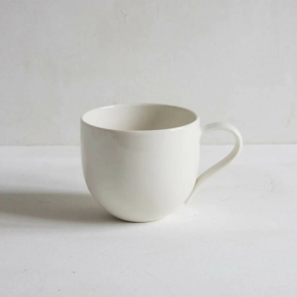 A simple, off-white porcelain mug with a handle, placed against a neutral background , available  to buy on the-salon.com. SALON curates the finest , unique, handmade pieces from the interior industry's best makers and brands. Shop luxury furniture, lighting, rugs, art, antiques and vintage pieces and accessories from SALON interior designed rooms.