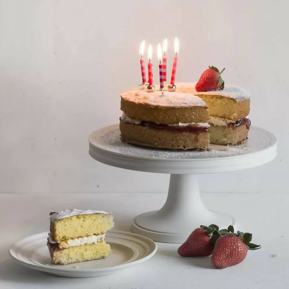 A white porcelain cake stand with a classic design, featuring a round platform on a cylindrical pedestal, with a Victoria sponge cake,   available  to buy on the-salon.com. SALON curates the finest , unique, handmade pieces from the interior industry's best makers and brands. Shop luxury furniture, lighting, rugs, art, antiques and vintage pieces and accessories from SALON interior designed rooms.