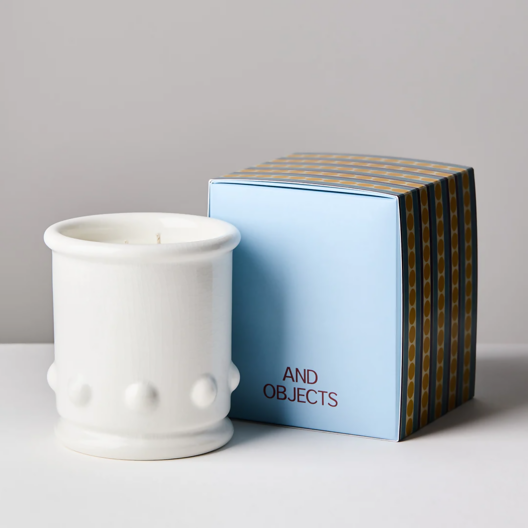 A white ceramic candle vessel with spherical detailing on the sides, with a candle visible at the top, with the box next to it. The vessel is placed against a neutral background,  available  to buy on the-salon.com. SALON curates the finest , unique, handmade pieces from the interior industry's best makers and brands. Shop luxury furniture, lighting, rugs, art, antiques and vintage pieces and accessories from SALON interior designed rooms.