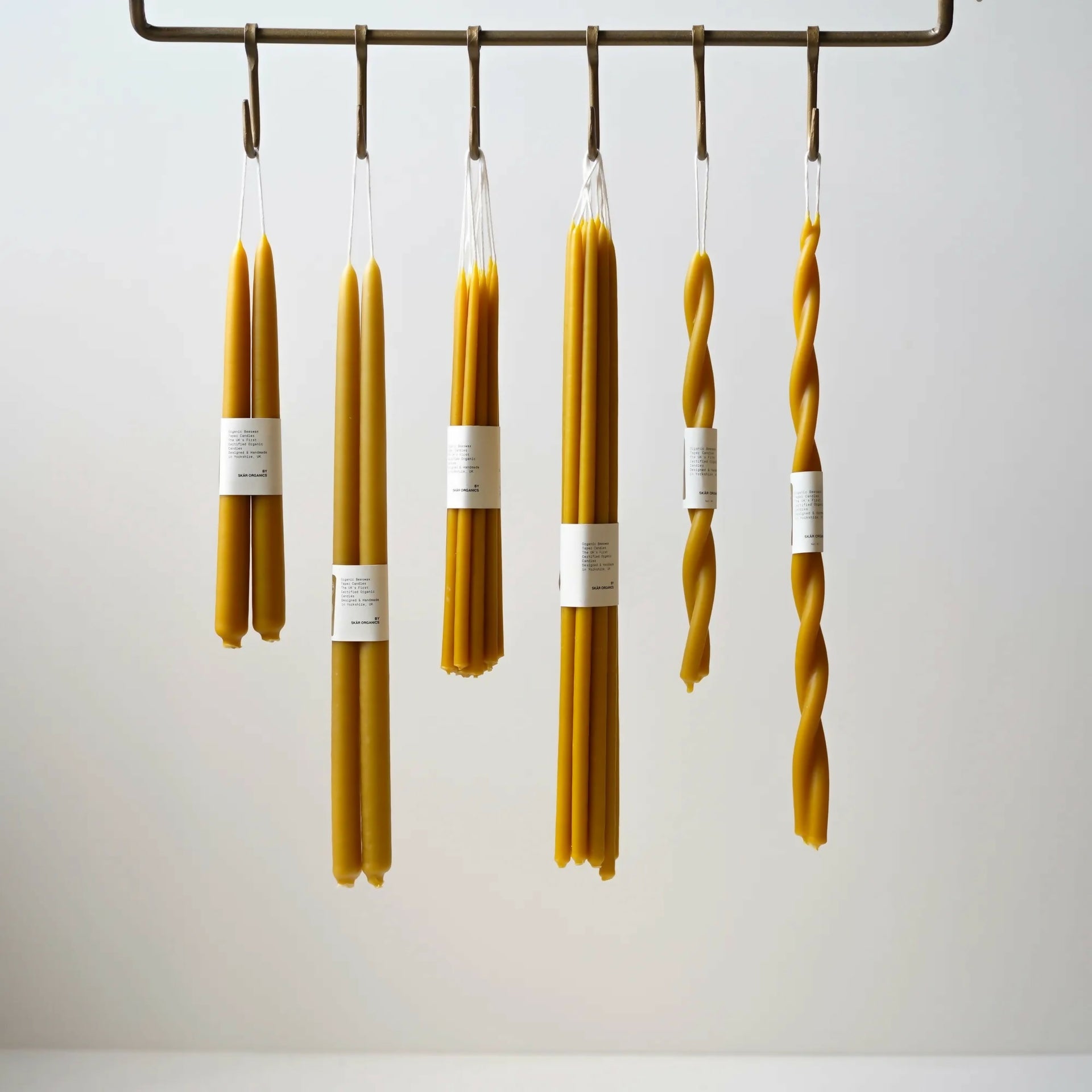 A group of unlit organic beeswax dinner candles, in various shapes including straight and twisted, hanging from a metal rod,  available  to buy on the-salon.com. SALON curates the finest , unique, handmade pieces from the interior industry's best makers and brands. Shop luxury furniture, lighting, rugs, art, antiques and vintage pieces and accessories from SALON interior designed rooms.