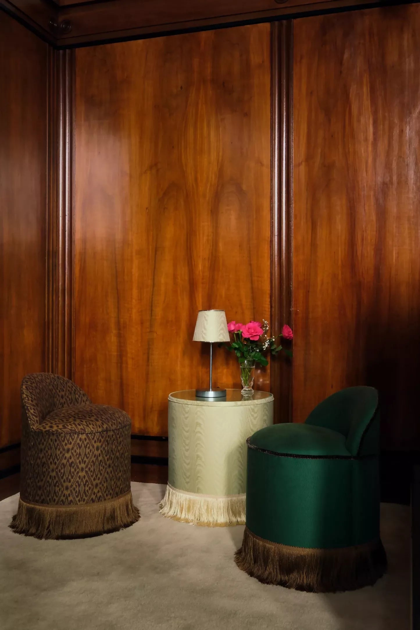 A green upholstered Inferi stool with a curvilinear backrest and a brown fringe at the base, in a room with hardwood walls and another unholstered chair, designed for room service or salon use,  available  to buy on the-salon.com. SALON curates the finest , unique, handmade pieces from the interior industry's best makers and brands. Shop luxury furniture, lighting, rugs, art, antiques and vintage pieces and accessories from SALON interior designed rooms.