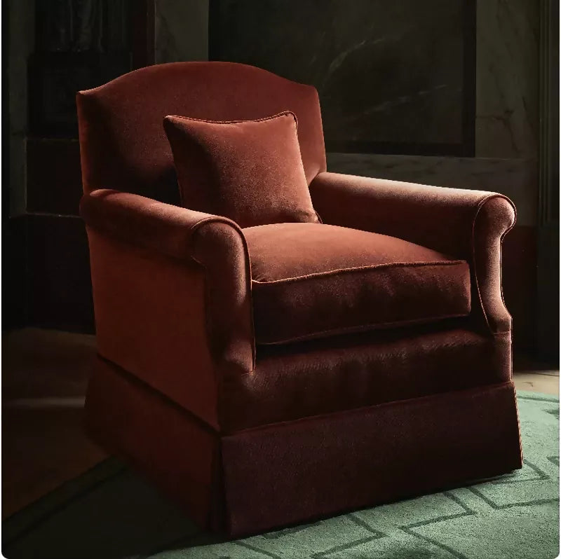 A brown upholstered armchair with classic curved arms and a plump cushion, in a room with a dark background, a grey carpet and liminal lighting,  available  to buy on the-salon.com. SALON curates the finest , unique, handmade pieces from the interior industry's best makers and brands. Shop luxury furniture, lighting, rugs, art, antiques and vintage pieces and accessories from SALON interior designed rooms.