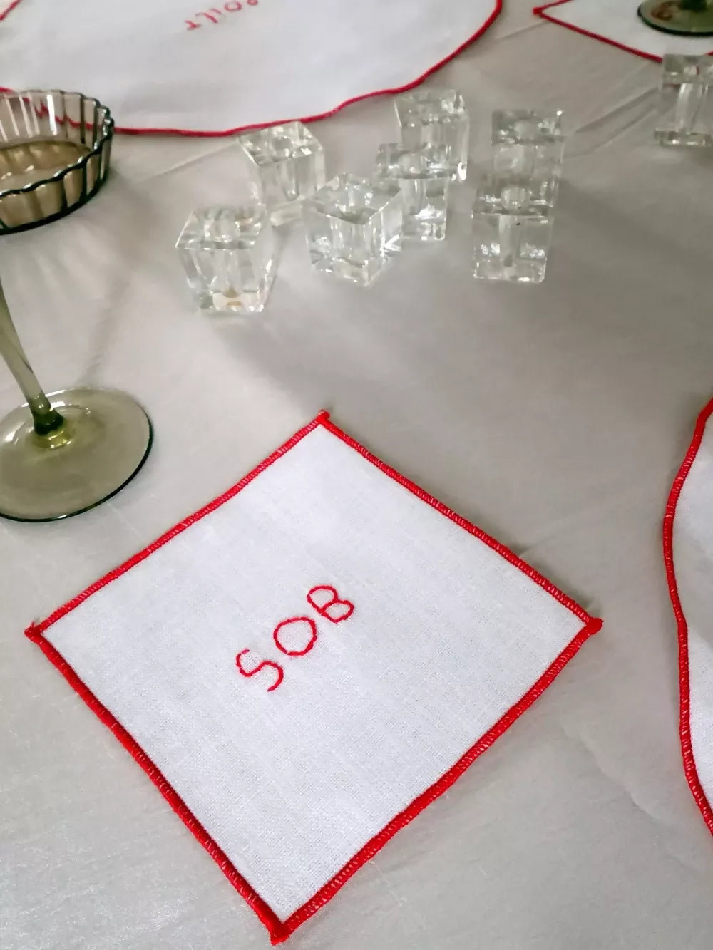White napkin with 'SOB' embroidered on it, placed on a table with glasses and a glass holder, available  to buy on the-salon.com. SALON curates the finest , unique, handmade pieces from the interior industry's best makers and brands. Shop luxury furniture, lighting, rugs, art, antiques and vintage pieces and accessories from SALON interior designed rooms.