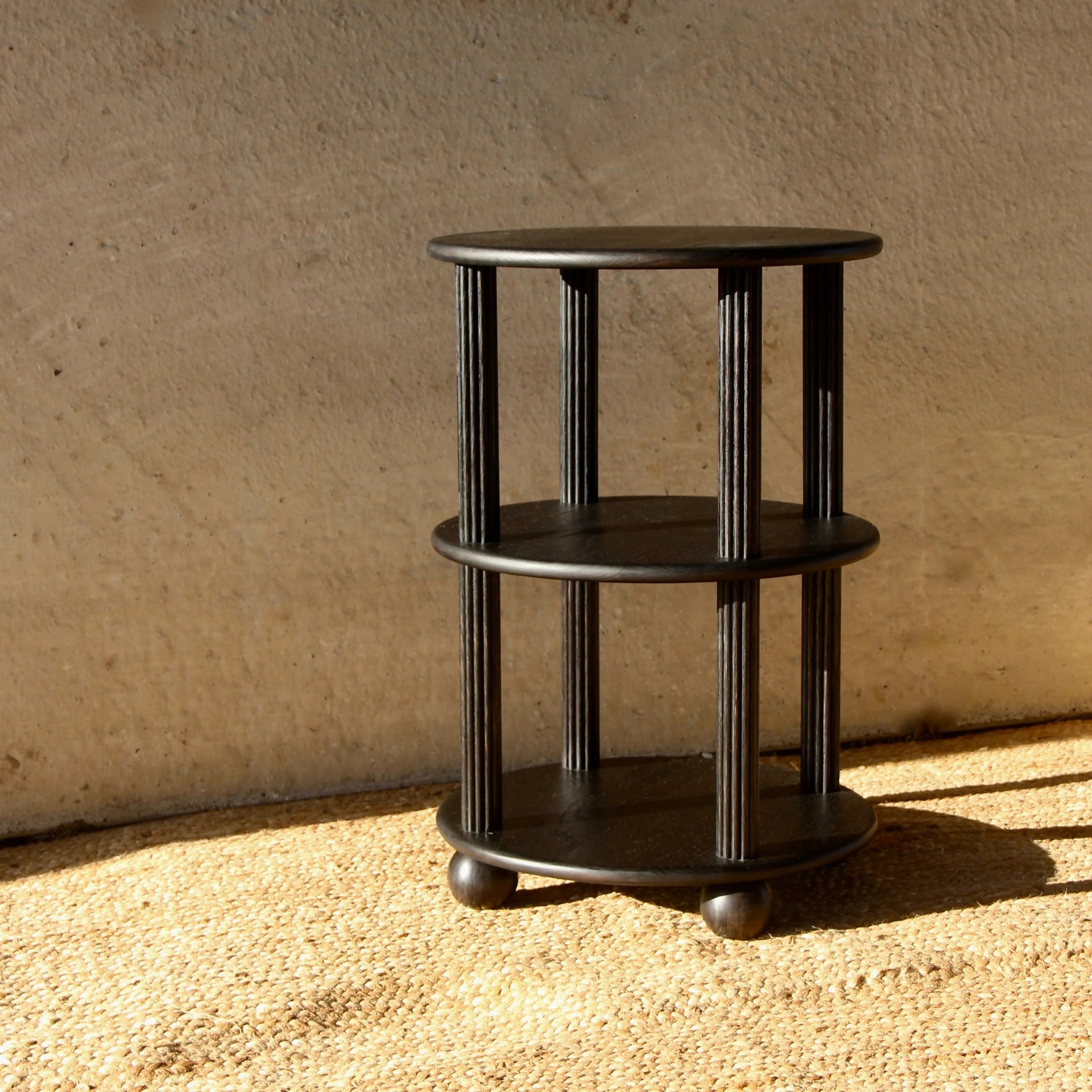 A black, three-tiered oak what-not table with reeded pillars and rounded bun feet, placed on a textured surface with a shadow cast across its legs,  available  to buy on the-salon.com. SALON curates the finest , unique, handmade pieces from the interior industry's best makers and brands. Shop luxury furniture, lighting, rugs, art, antiques and vintage pieces and accessories from SALON interior designed rooms.