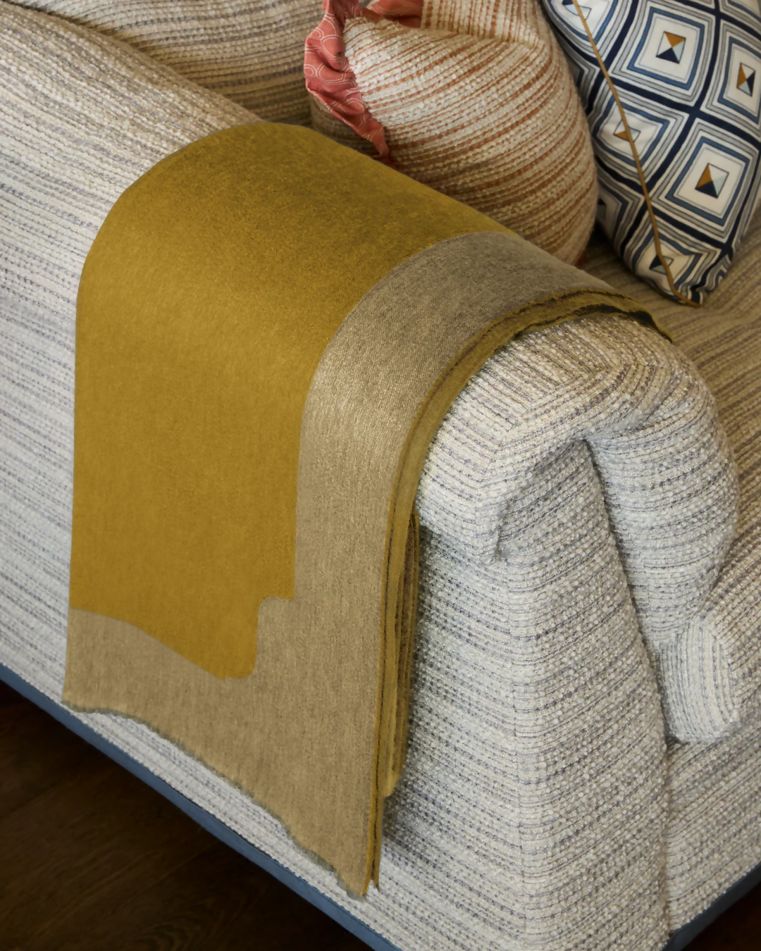 A gold-coloured cashmere throw blanket with a decorative border, displaying a small blue and silver brand tag on the bottom right corner,  available  to buy on the-salon.com. SALON curates the finest , unique, handmade pieces from the interior industry's best makers and brands. Shop luxury furniture, lighting, rugs, art, antiques and vintage pieces and accessories from SALON interior designed rooms.