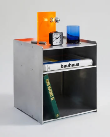 A minimalist silver metal side table with the text 'bauhaus' on it, featuring a small clock, an orange vase, and a blue glass on top, with a green book visible inside the table's space,  available  to buy on the-salon.com. SALON curates the finest , unique, handmade pieces from the interior industry's best makers and brands. Shop luxury furniture, lighting, rugs, art, antiques and vintage pieces and accessories from SALON interior designed rooms.