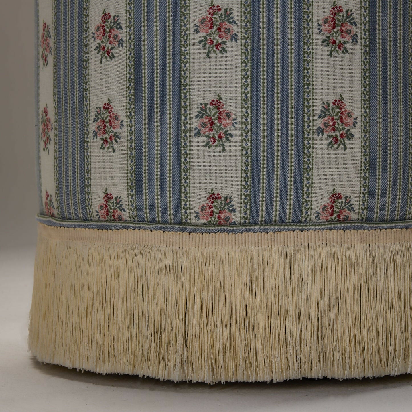 A close up of the round footstool upholstered in a multicoloured fabric with stripes and floral patterns, featuring a silky champagne fringe at the bottom,  available  to buy on the-salon.com. SALON curates the finest , unique, handmade pieces from the interior industry's best makers and brands. Shop luxury furniture, lighting, rugs, art, antiques and vintage pieces and accessories from SALON interior designed rooms.