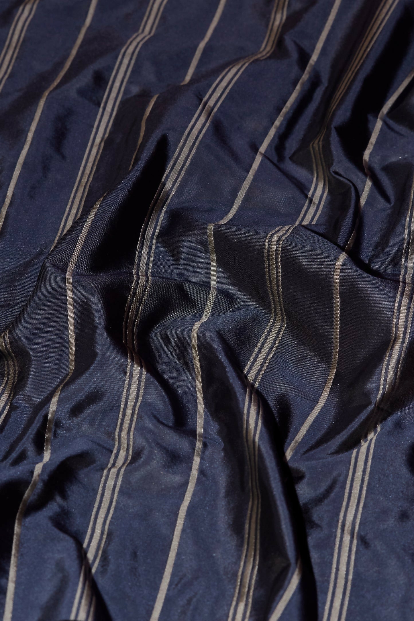 Close-up view of tafta stripe fabric in navy with a contrasting silver stripe, showcasing its texture and reflective quality,  available  to buy on the-salon.com. SALON curates the finest , unique, handmade pieces from the interior industry's best makers and brands. Shop luxury furniture, lighting, rugs, art, antiques and vintage pieces and accessories from SALON interior designed rooms.