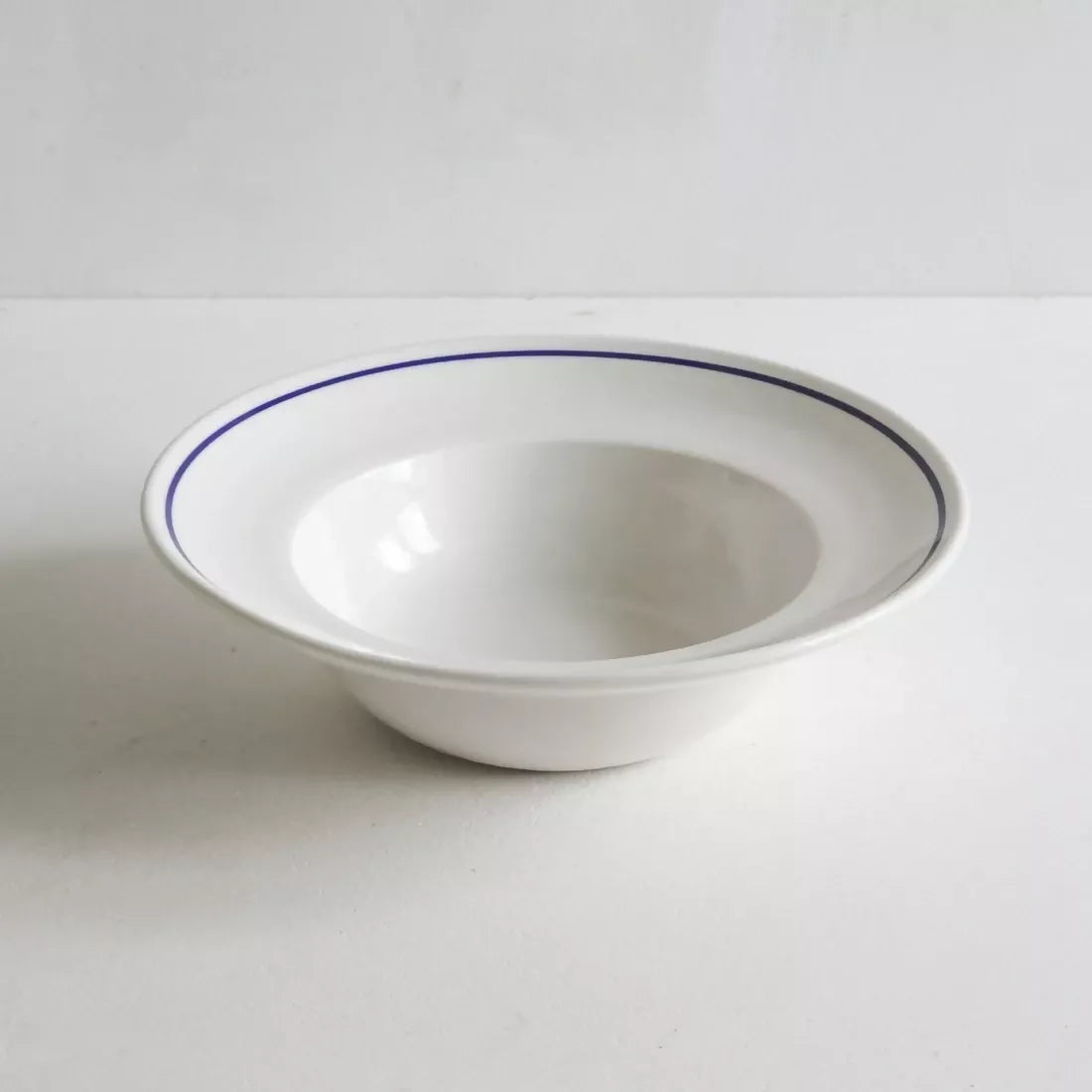 A white porcelain deep bowl with a classic cobalt blue line along the rim, against a plain background, available  to buy on the-salon.com. SALON curates the finest , unique, handmade pieces from the interior industry's best makers and brands. Shop luxury furniture, lighting, rugs, art, antiques and vintage pieces and accessories from SALON interior designed rooms.