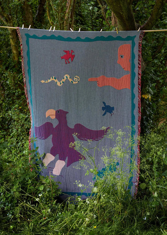 A colorful handcrafted blanket with animal and imaginative figures displayed infront of trees and a woody background, titled 'To ASAFO PARK THROW BLANKET BY AMECHI',  available  to buy on the-salon.com. SALON curates the finest , unique, handmade pieces from the interior industry's best makers and brands. Shop luxury furniture, lighting, rugs, art, antiques and vintage pieces and accessories from SALON interior designed rooms.