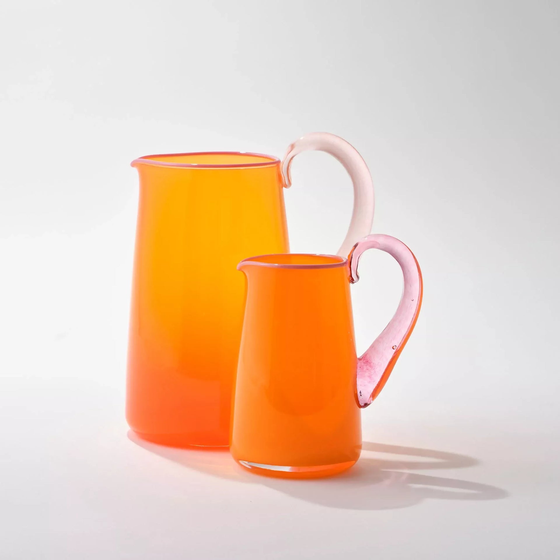 Two hand blown, transparent orange glass jugs with a blush handle and contrasting pale pink lips, one in a smaller size- on a white background,  available  to buy on the-salon.com. SALON curates the finest , unique, handmade pieces from the interior industry's best makers and brands. Shop luxury furniture, lighting, rugs, art, antiques and vintage pieces and accessories from SALON interior designed rooms.