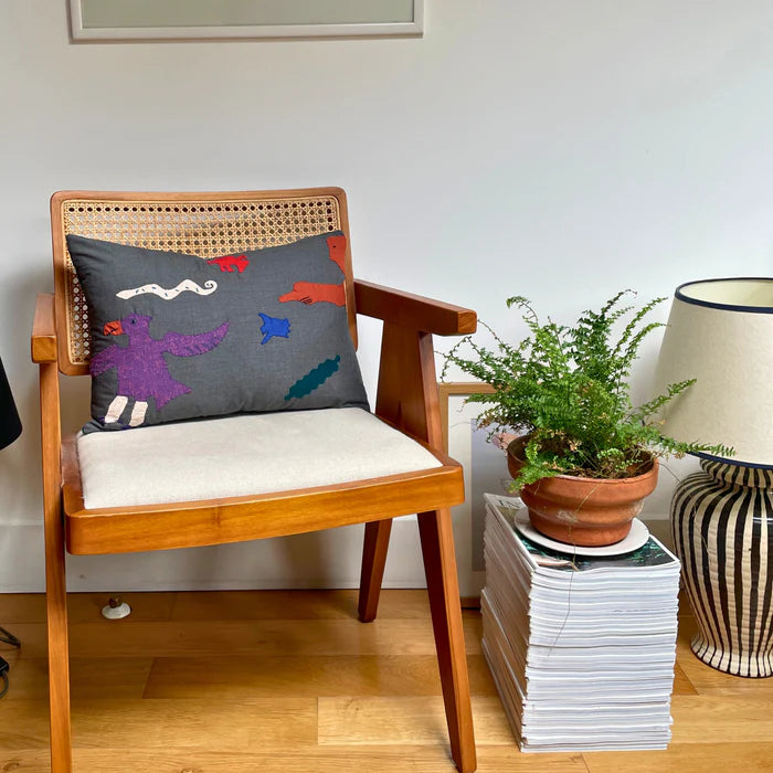 A canvas cushion with a colourful character design placed on a wooden chair, with a potted plant and lamp in the background,  available  to buy on the-salon.com. SALON curates the finest , unique, handmade pieces from the interior industry's best makers and brands. Shop luxury furniture, lighting, rugs, art, antiques and vintage pieces and accessories from SALON interior designed rooms.