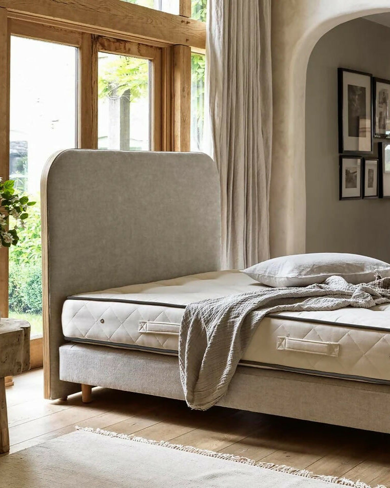 Beige upholstered bed with a textured headboard in a room with large windows and neutral decor,  available  to buy on the-salon.com. SALON curates the finest , unique, handmade pieces from the interior industry's best makers and brands. Shop luxury furniture, lighting, rugs, art, antiques and vintage pieces and accessories from SALON interior designed rooms.