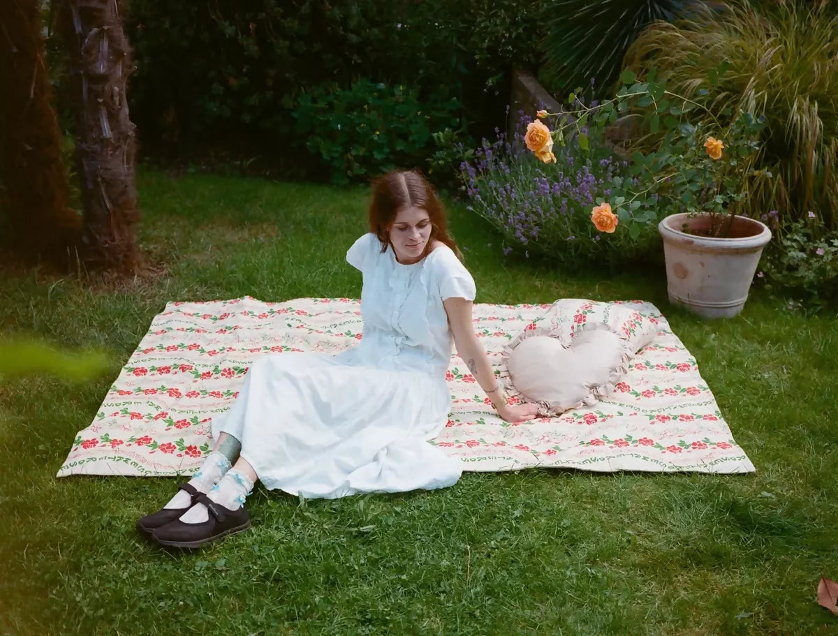 A quilt with a floral pattern featuring roses in red and green colours on grass with a woman in a white dress on it,  available  to buy on the-salon.com. SALON curates the finest , unique, handmade pieces from the interior industry's best makers and brands. Shop luxury furniture, lighting, rugs, art, antiques and vintage pieces and accessories from SALON interior designed rooms.