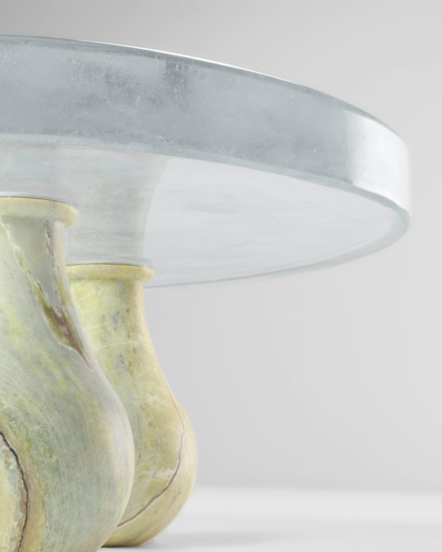 A close-up studio shot of a round table with a thick brushed silver metal top and three sculptural legs made of pale marbled stone in cream and yellow-green tones.