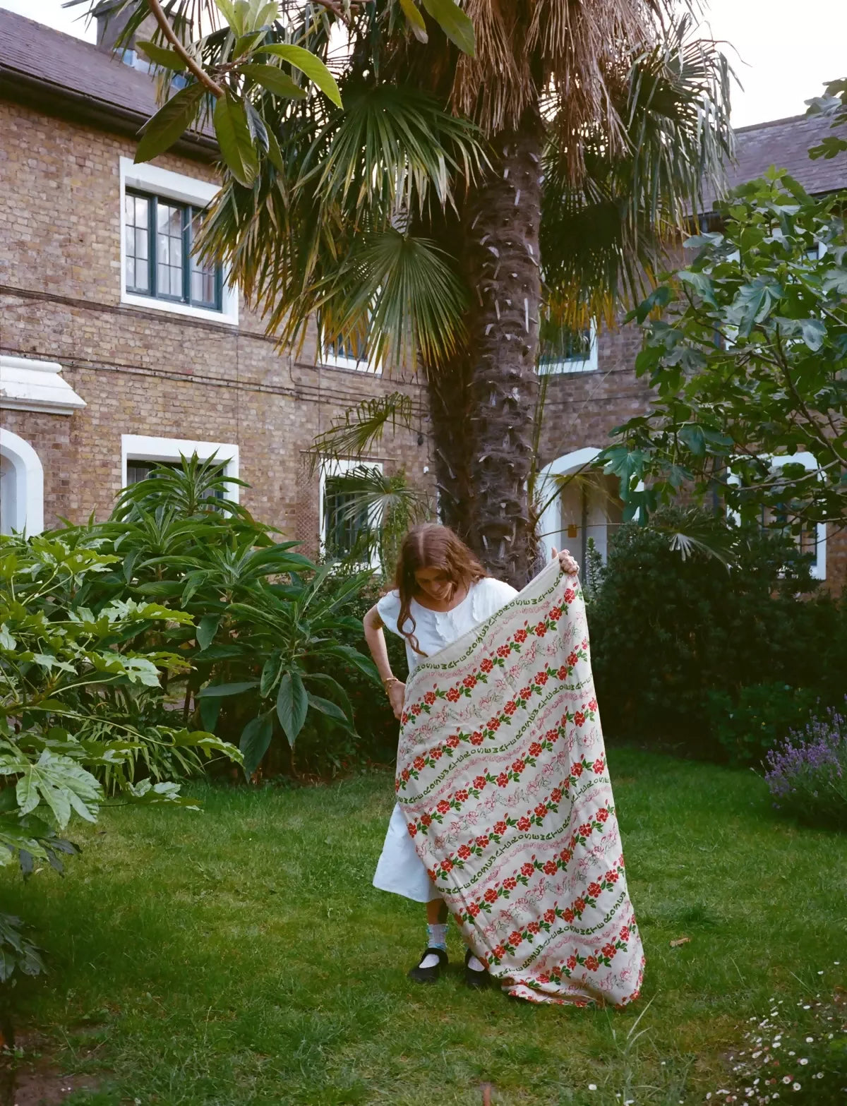 A quilt with a floral pattern featuring roses in red and green colors with a woman holding it outside ,  available  to buy on the-salon.com. SALON curates the finest , unique, handmade pieces from the interior industry's best makers and brands. Shop luxury furniture, lighting, rugs, art, antiques and vintage pieces and accessories from SALON interior designed rooms.
