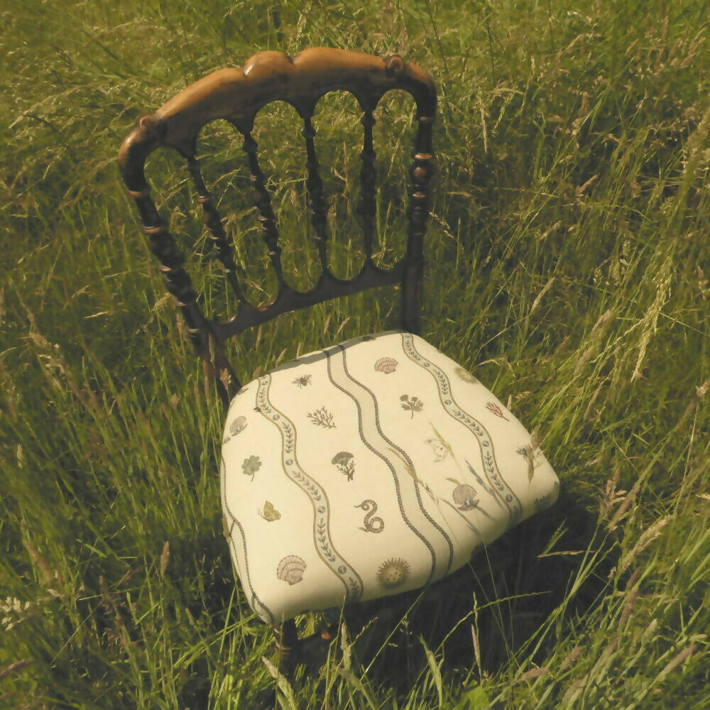 A detailed image of a fabric with a natural and whimsical pattern design, featuring elements such as a snake, a sun, a shell, a clover, and a butterfly, with a moon and leaf motifs,  available  to buy on the-salon.com. SALON curates the finest , unique, handmade pieces from the interior industry's best makers and brands. Shop luxury furniture, lighting, rugs, art, antiques and vintage pieces and accessories from SALON interior designed rooms.