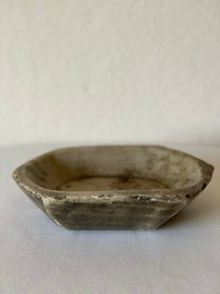 A primitive carved marble bowl with a hexagonal shape, predominantly gray in color, showcasing natural stone patterns, available  to buy on the-salon.com. SALON curates the finest , unique, handmade pieces from the interior industry's best makers and brands. Shop luxury furniture, lighting, rugs, art, antiques and vintage pieces and accessories from SALON interior designed rooms.