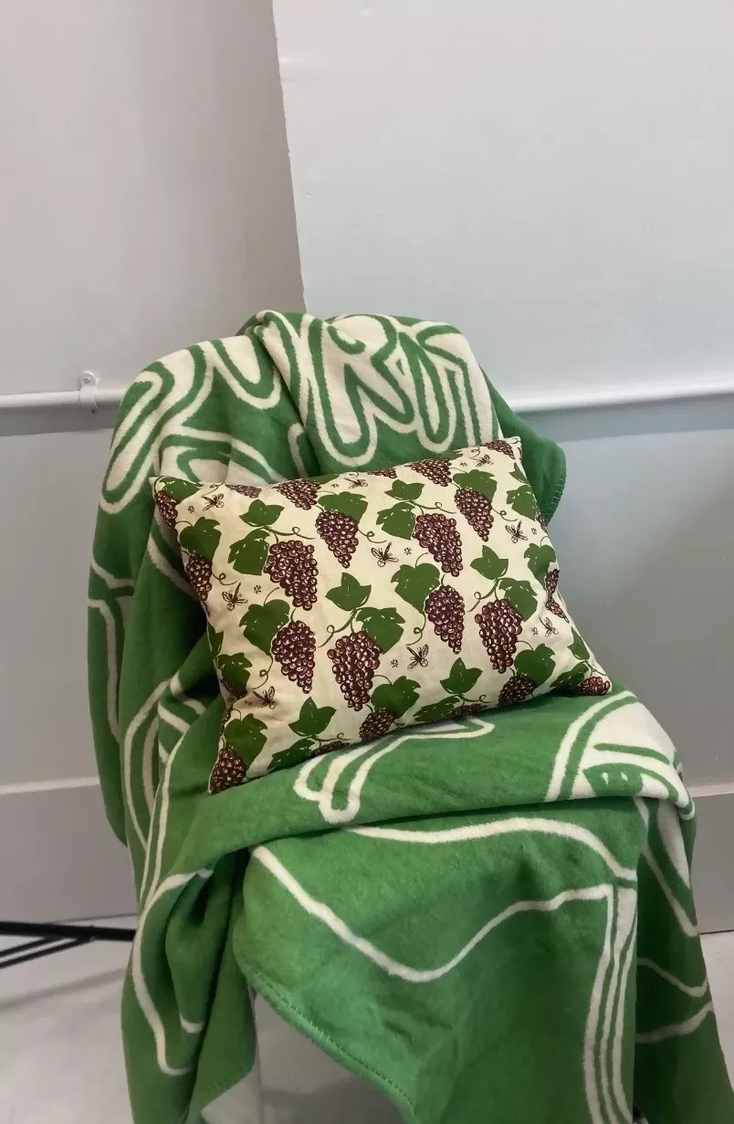 A rectangular cushion with a floral pattern featuring purple grapes and green leaves, placed on a green textile with a leaf pattern,  available  to buy on the-salon.com. SALON curates the finest , unique, handmade pieces from the interior industry's best makers and brands. Shop luxury furniture, lighting, rugs, art, antiques and vintage pieces and accessories from SALON interior designed rooms.
