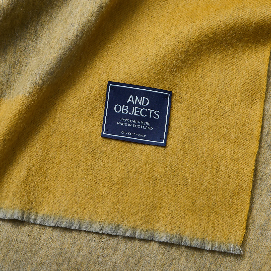 A folded gold-colored cashmere throw blanket with a decorative border, displaying a small blue and silver brand tag on the bottom right corner,  available  to buy on the-salon.com. SALON curates the finest , unique, handmade pieces from the interior industry's best makers and brands. Shop luxury furniture, lighting, rugs, art, antiques and vintage pieces and accessories from SALON interior designed rooms.